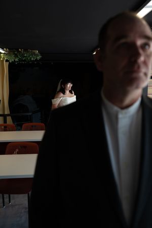 Groom in black suit stands in foreground as bride in white off-shoulder gown waits in dimly lit venue background.