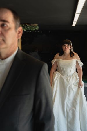 Bride in elegant off-shoulder white wedding gown stands behind man in gray suit in dimly lit venue.
