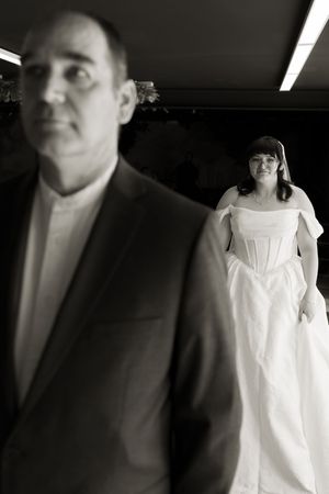 Black and white photo of bride in white gown standing behind man in suit jacket indoors.