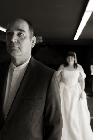 Black and white dramatic portrait of man in suit with bride in white gown in background.