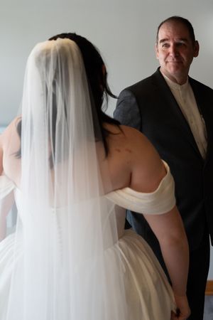A bride in a white off-shoulder gown and veil faces a smiling man in a black suit indoors.