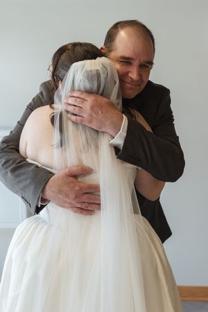 Father of the bride sharing emotional hug with bride in white wedding gown and veil indoors.