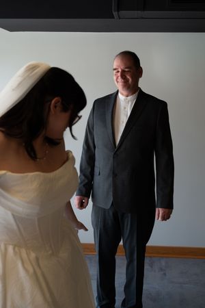 Groom in black suit smiles at bride in white wedding dress during indoor first look.