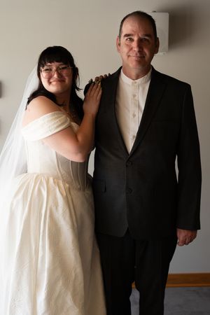 Bride in white off-shoulder gown with veil poses with groom in black suit indoors.