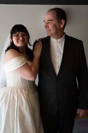 Smiling bride in white wedding gown and veil stands beside groom in black suit indoors.