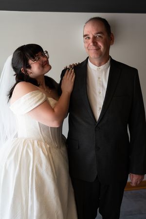 Bride in white off-shoulder dress with veil rests hand on groom's shoulder at wedding.
