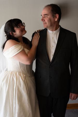 Bride in white off-shoulder gown and veil embraces groom in black suit indoors.