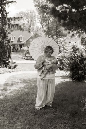Black and white photo of a woman holding a newborn baby under a white parasol in a garden.