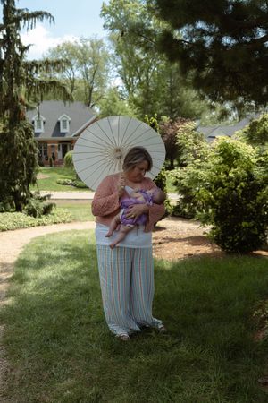 Woman holding baby and white parasol umbrella in lush garden setting on sunny day.