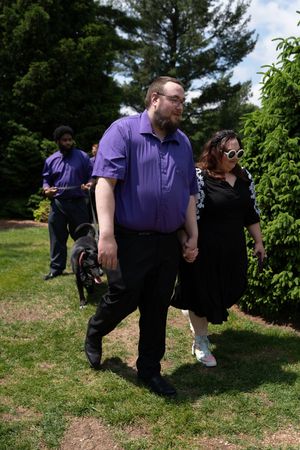 Couple holding hands walking through garden, man in purple shirt and woman in black dress with white sunglasses.