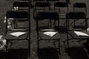 Black and white photo of folding chairs with paper fans on seats set up for outdoor ceremony.