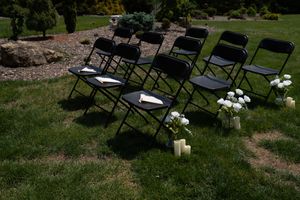 Black folding chairs arranged on green grass with white roses and candles for outdoor wedding.