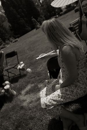 Black and white photo of blonde woman in floral dress holding fan at outdoor wedding ceremony on sunny day.