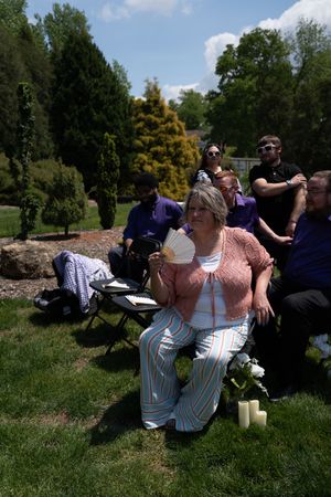 Woman in pink top and striped pants fans herself at an outdoor garden gathering with guests seated on a sunny day.