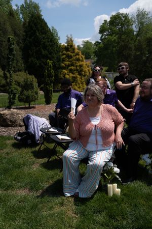 Woman in pink top and striped pants fans herself while seated at sunny outdoor wedding ceremony with guests behind her.