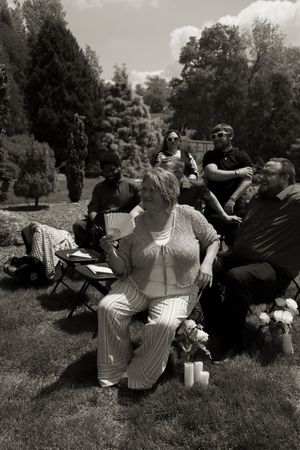 Black and white photo of wedding guests seated outdoors, woman with fan in foreground, trees and shrubs in background.