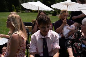 Young male wedding guest in pink shirt seated among guests at sunny outdoor ceremony.