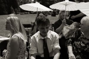 Black and white photo of wedding guests sitting outdoors under white parasols at a garden ceremony.