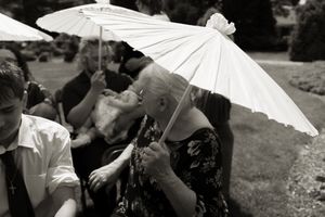 Black and white photo of wedding guests shading themselves with white parasols at outdoor ceremony.