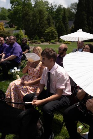 Wedding guests seated outdoors using parasols and fans to shade from summer sun.