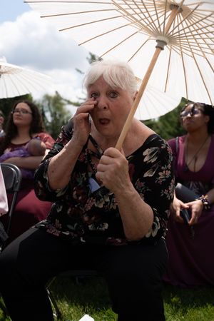 Emotional elderly woman holding white parasol wipes tear during outdoor wedding ceremony.