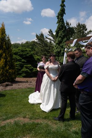 Bride in white gown stands at outdoor wedding ceremony surrounded by lush green trees.