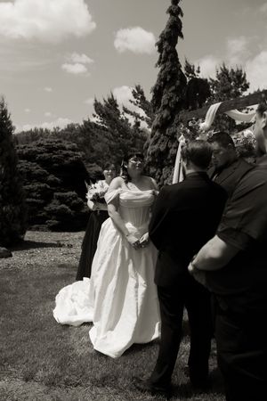 Black and white photo of a bride in a white gown holding bouquet during an outdoor wedding ceremony.