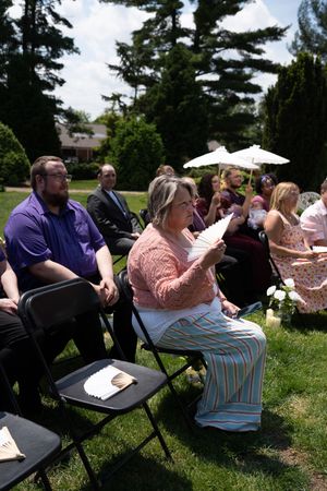 Wedding guests seated in black folding chairs outdoors, with a woman fanning herself in the foreground.