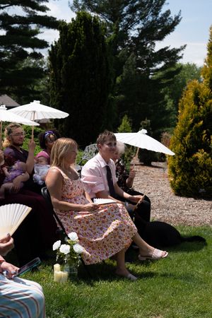 Wedding guests seated outdoors using white parasols and fans to shade from sun during garden ceremony on bright day.