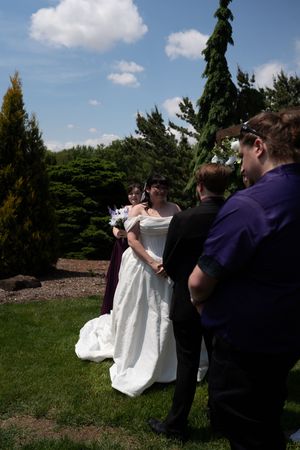 Bride in white strapless gown holds purple bouquet during outdoor wedding ceremony surrounded by guests and evergreen trees.