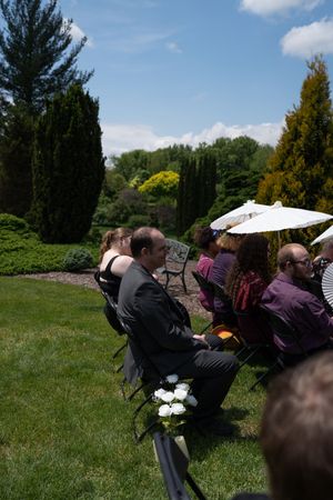 Guests seated in chairs on green lawn during sunny outdoor wedding ceremony, white parasol visible among attendees.