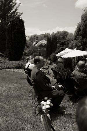 Wedding guests seated on chairs in a lush garden during an outdoor ceremony, one holding a parasol.