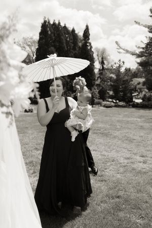 Black and white photo of a bridesmaid in a black dress holding a baby and white parasol at an outdoor wedding.