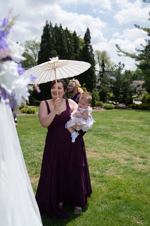 Bridesmaid in a burgundy gown holding a white parasol and a baby at an outdoor wedding ceremony.