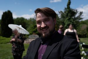 Smiling bearded man in black suit and purple shirt at sunny outdoor wedding celebration.