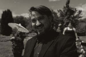 Smiling man in black suit at outdoor wedding ceremony with guests and parasols in background.