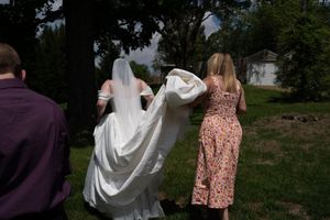 A bridesmaid in a floral dress helps carry a bride's white wedding gown and veil outdoors.