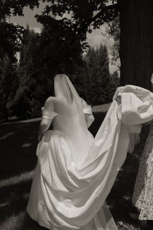 Bride in white off-shoulder gown and veil walks through garden in black and white photo.
