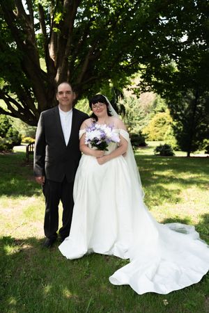 Bride in white gown holding purple bouquet poses with groom in dark suit outdoors in lush green park.