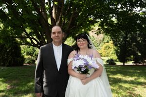 Bride and groom pose together outdoors under green trees, bride holding purple and white bouquet.