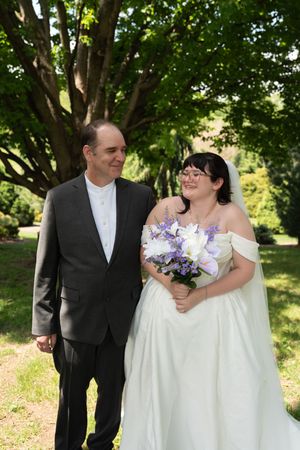 A bride in a white gown holding purple flowers stands smiling beside a man in a gray suit outdoors.