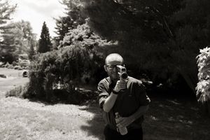 Black and white photo of bald man holding vintage camera outdoors in lush garden setting.