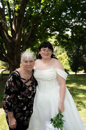Bride in white off-shoulder gown holds bouquet beside older woman in floral dress outdoors.