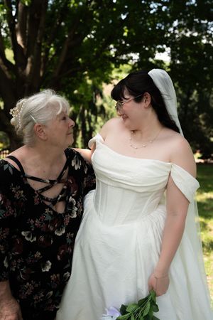 Elderly woman admiring bride in white off-shoulder wedding gown with veil outdoors.