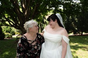 An older woman and a bride in a white off-shoulder gown share a warm smile outdoors under trees.