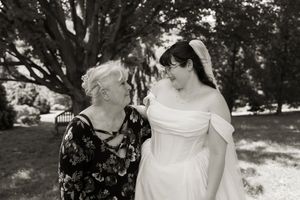 Black and white photo of bride in white off-shoulder gown smiling with older woman outdoors.