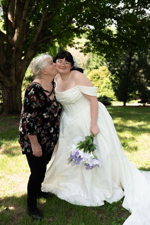 Older woman kisses bride in white off-shoulder wedding gown on cheek while standing on green lawn under trees.