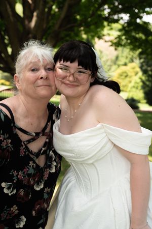 Bride in white off-shoulder gown smiles with older woman in floral dress at outdoor wedding celebration.