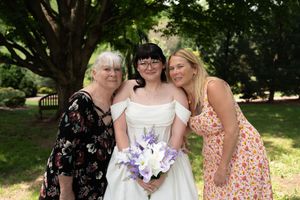 Bride in white off-shoulder gown holding purple flowers posing with two women outdoors.