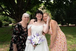 Bride in white gown holding purple bouquet posing with two women under trees in garden.